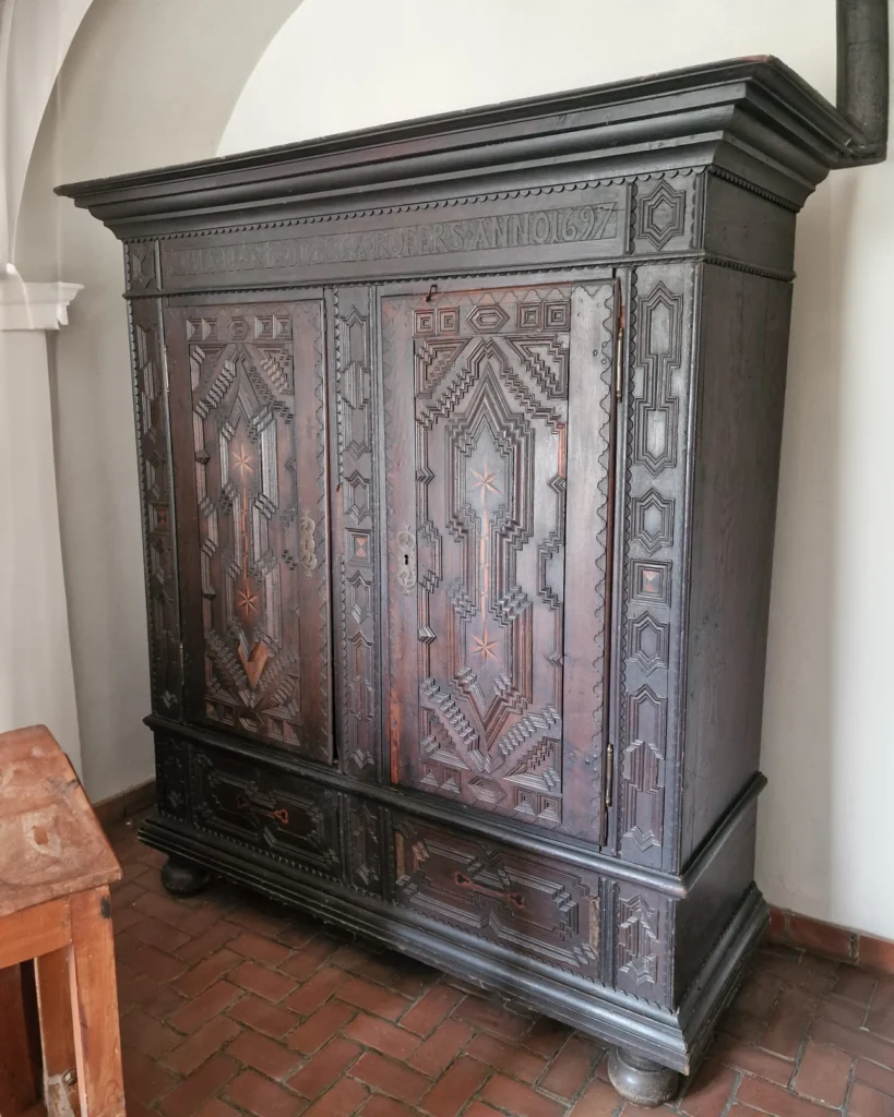 Stara, ciemna drewniana szafa z bogato rzeźbionymi geometrycznymi zdobieniami i napisem datowanym na 1627 rok. Mebel stoi na ceglanej podłodze we wnętrzu o łukowym sklepieniu.