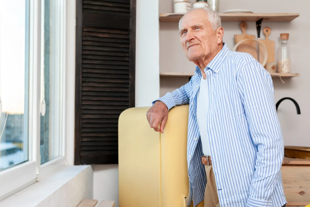 Starszy mężczyzna stojący w kuchni, oparty o żółtą lodówkę. Ubrany jest w jasne spodnie, biały podkoszulek i koszulę w niebiesko-białe paski. Patrzy przez okno z zamyśleniem. W tle widoczne są drewniane półki z kuchennymi akcesoriami.