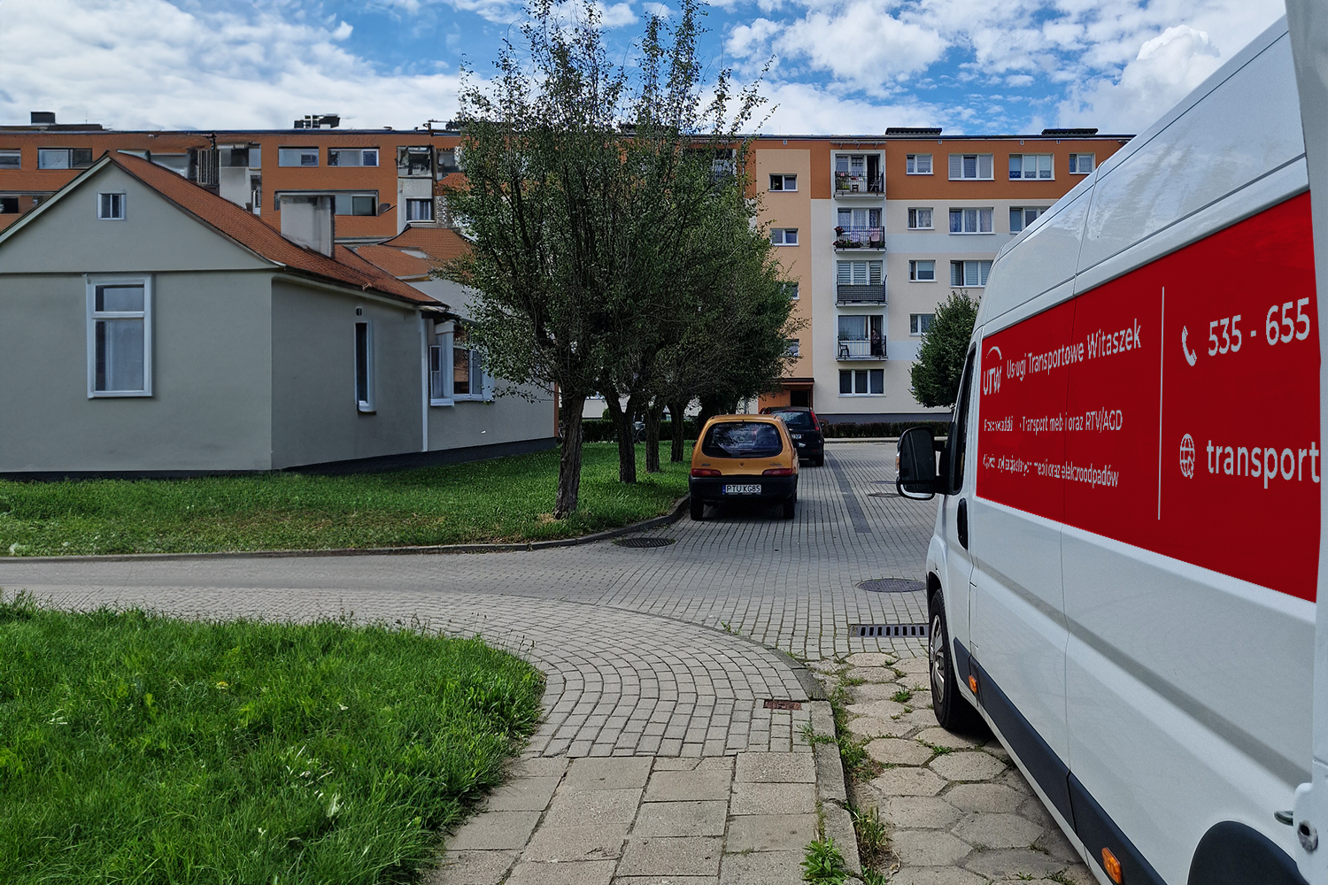 Biała furgonetka firmy Usługi Transportowe Witaszek z czerwonym panelem reklamowym zaparkowana na osiedlu. Na panelu widnieje numer telefonu i oferta usług: przeprowadzki, transport mebli i sprzętu RTV/AGD oraz wywóz odpadów. W tle widać niski domek z czerwonym dachem, drzewa oraz blok mieszkalny z balkonami.