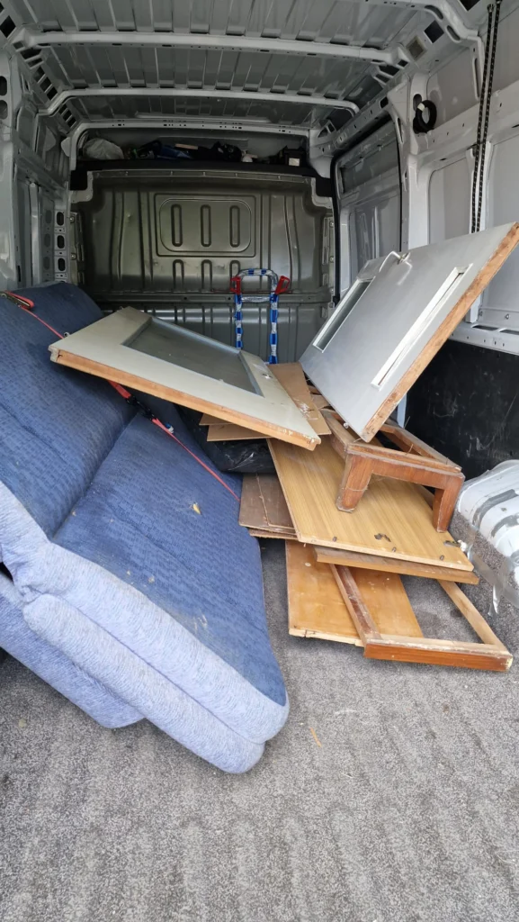 Wnętrze furgonetki wypełnione starymi meblami przeznaczonymi do utylizacji. W środku znajduje się złożona niebieska kanapa, drewniane drzwi, fragmenty szafek oraz inne elementy drewniane ułożone luzem.